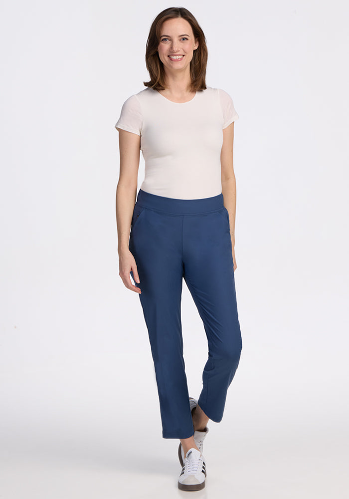 A woman with straight, shoulder-length brown hair poses casually against a white background, wearing a white tee, Woolx Nicole Petite Ankle Pant in Vintage Indigo, and white sneakers. She smiles warmly, showcasing a relaxed style.