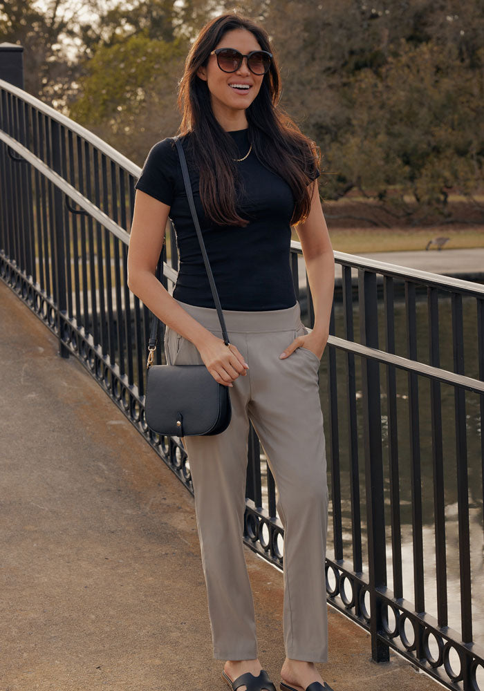 A woman with long dark hair and sunglasses, wearing a black t-shirt, Woolx Nicole Petite Ankle Pant in Vintage Khaki, black slip-on shoes, and a black crossbody bag stands on a bridge with one hand in her pocket, smiling outdoors.
