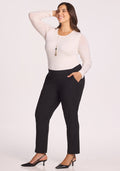 A woman wearing the Woolx Nicole Tall Ankle Pant in black, paired with a long-sleeve white top, black heels, and a long necklace, stands smiling with one hand on her hip and the other touching her hair against a plain light background.