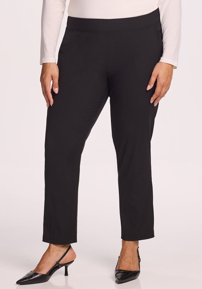 A person wears Woolx Nicole Tall Ankle Pant in black, paired with black low-heeled shoes and a light long-sleeve top. The image, cropped from the waist down, features a plain, light background.