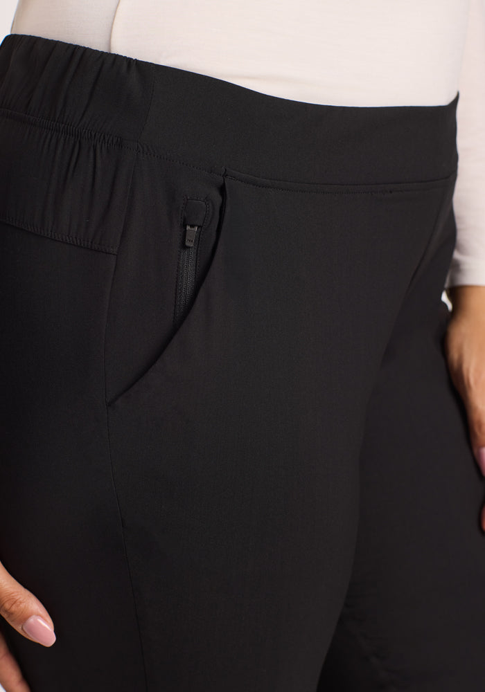 Close-up of a person wearing Woolx Nicole Tall Ankle Pant in black with tapered legs, elastic waistband, and zippered pocket, paired with a long-sleeve white top. These lightweight, breathable pants offer comfort as the hand rests near the pocket.