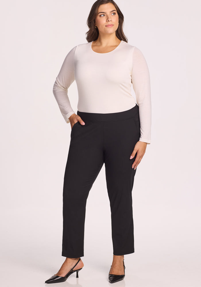 A woman with long brown hair wears a white long-sleeve top, black pointed-toe heels, and the Woolx Nicole Tall Ankle Pant in black. She stands with one hand in her pocket against a plain light background.