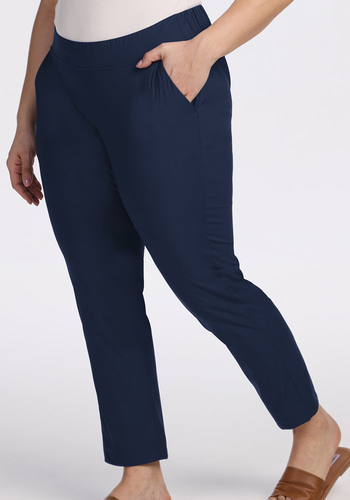 A person stands against a plain light background wearing Woolx Nicole Tall Ankle Pant in Deep Navy, paired with a white top and brown slip-on shoes. The pants are lightweight, breathable, and feature pockets.