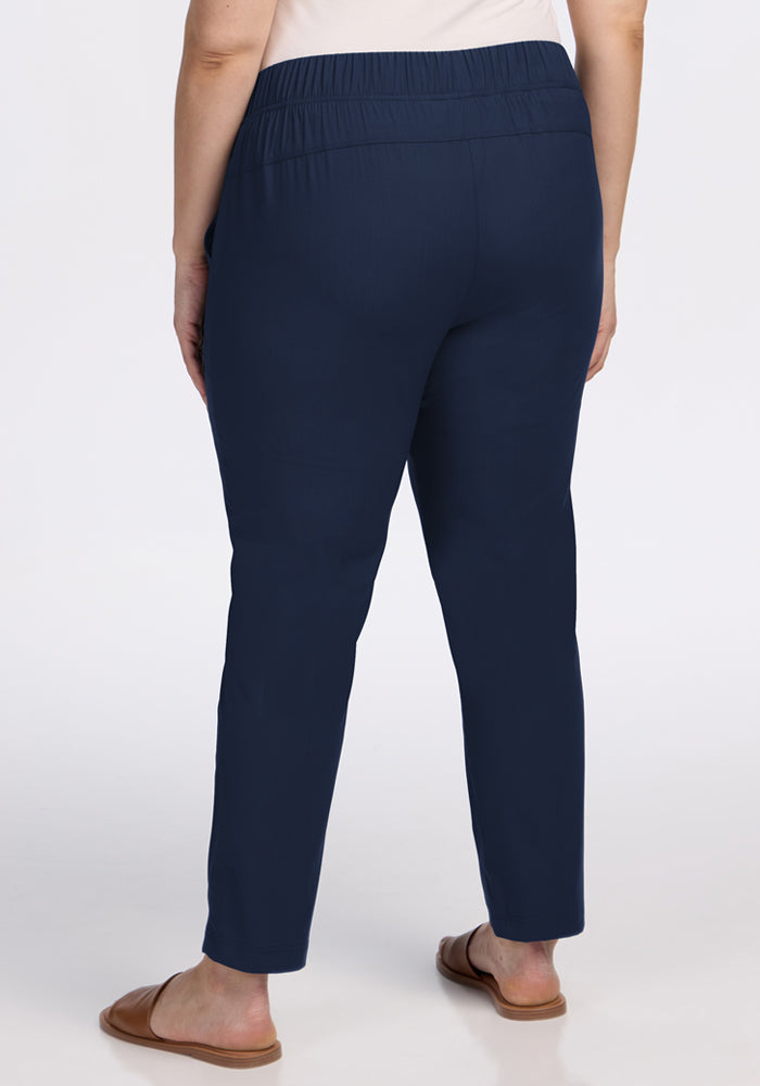 A person is shown from the waist down, wearing Woolx Nicole Tall Ankle Pant in Deep Navy and tan slip-on sandals, standing on a white background.