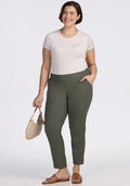 A woman smiles in a white short-sleeve top and Woolx Nicole Tall Ankle Pant in Sage Olive, holding a beige handbag. She pairs brown sandals and a necklace with her short brown hair. The background is plain white.
