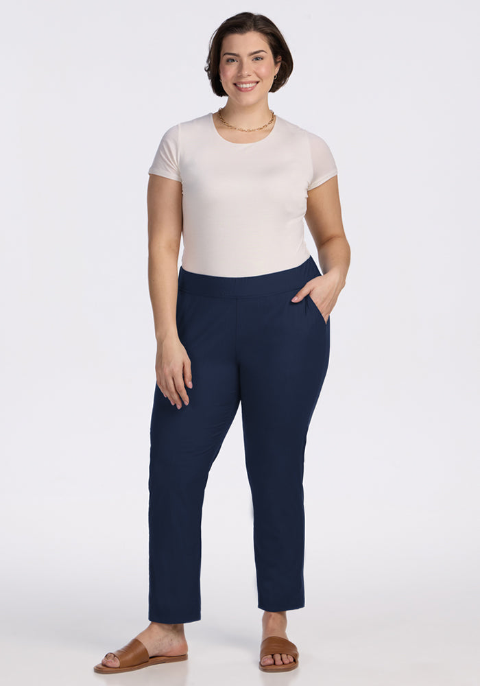 A woman with short brown hair models the Woolx Nicole Tall Ankle Pant in Deep Navy, paired with a fitted white t-shirt, brown sandals, and a necklace. She stands smiling with one hand in her pocket against a plain white background.
