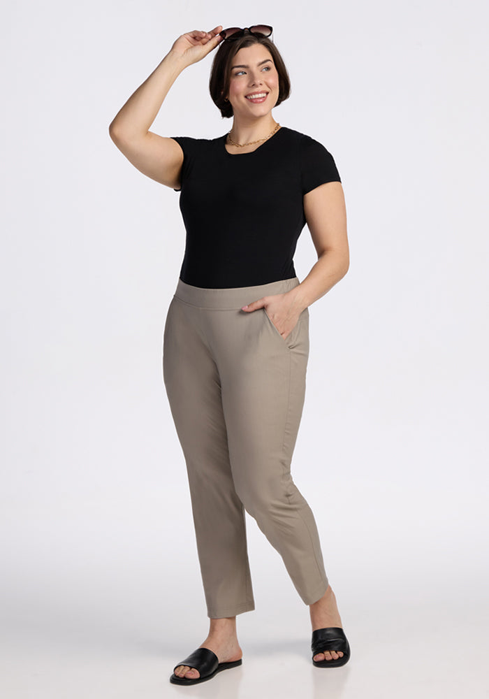 A woman stands smiling against a plain background, wearing a black t-shirt, Woolx Nicole Tall Ankle Pant in Vintage Khaki, black slide sandals, and sunglasses perched on her head—one hand on her hip, the other adjusting her shades.