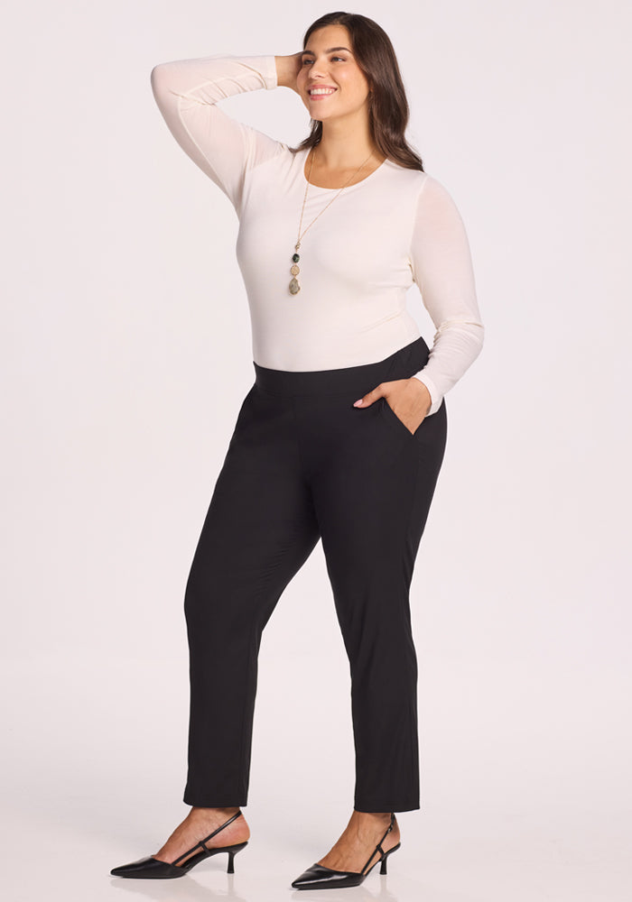 A woman wearing the Woolx Nicole Tall Ankle Pant in black, paired with a long-sleeve white top, black heels, and a long necklace, stands smiling with one hand on her hip and the other touching her hair against a plain light background.