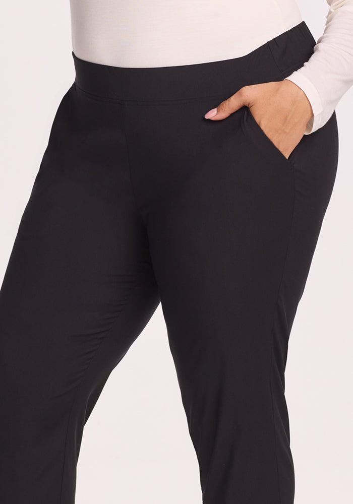 A person models the Woolx Nicole Tall Ankle Pant in black with a tapered-leg fit and a long-sleeve white top, standing with one hand in their pocket against a plain light background.