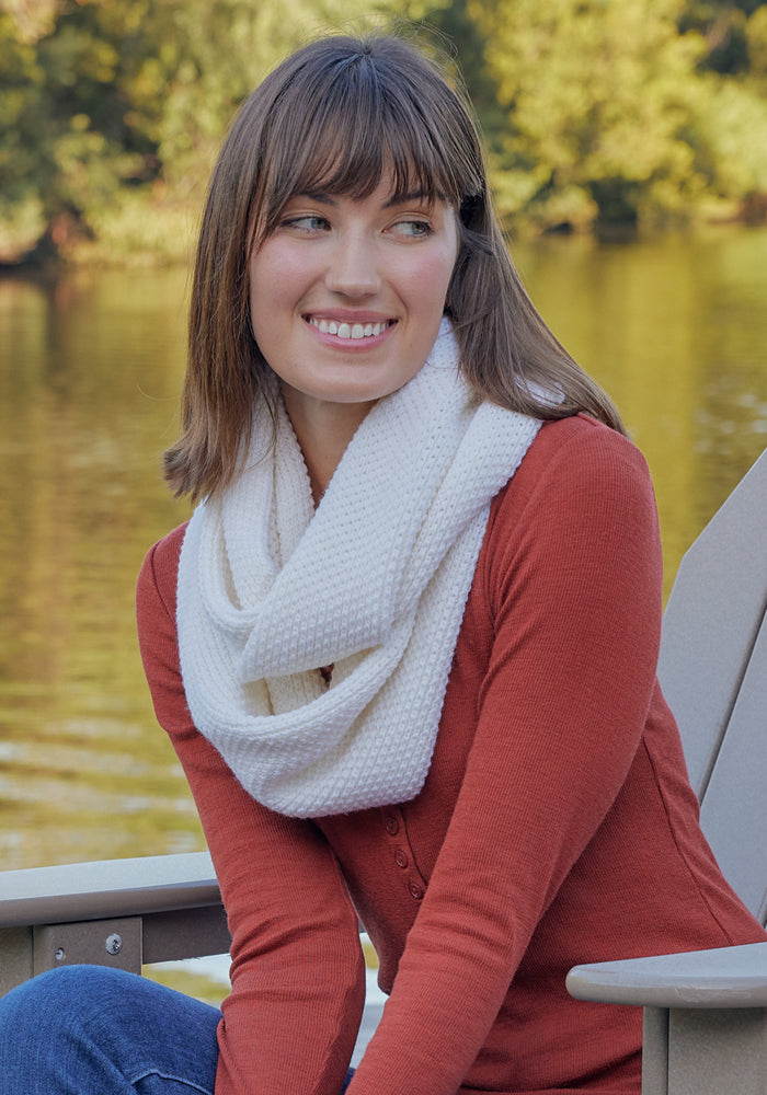 A woman with straight brown hair and bangs, wearing a rust-colored long-sleeve top and a Woolx Noelle Infinity Scarf in Buttercream, sits on a beige outdoor chair by the lake, smiling with trees in the background.