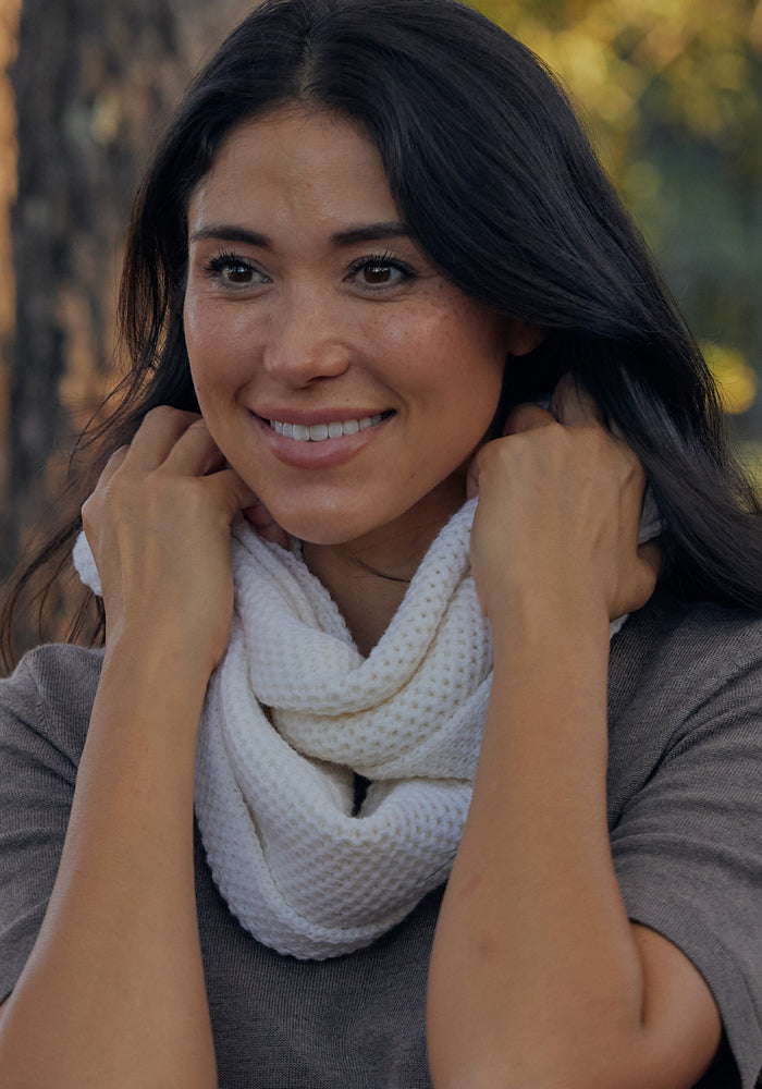 A woman with long dark hair smiles while adjusting the Woolx Noelle Infinity Scarf in Morning Fog around her neck. She is outdoors in a grey top, with sunlight and trees softly blurred in the background.