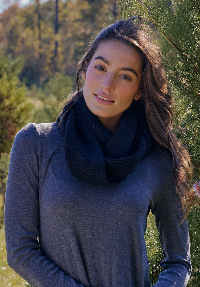 A woman with long brown hair, wearing the Woolx Noelle Infinity Scarf in black and a grey long-sleeve shirt, stands outdoors among green trees, smiling softly at the camera.