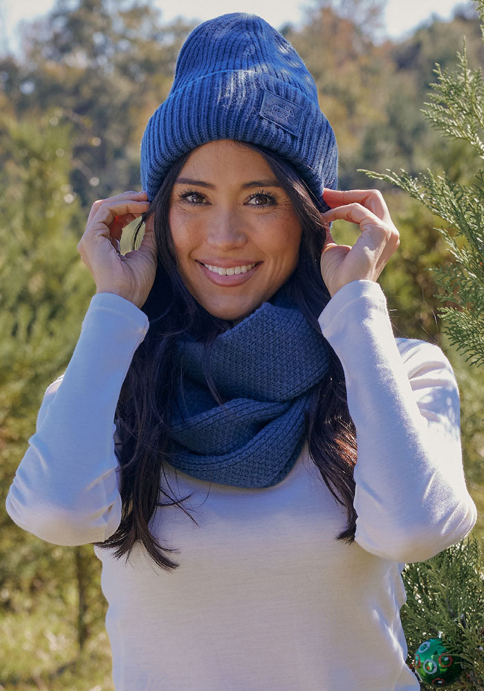 A woman with long dark hair wears a blue knit beanie and the Woolx Noelle Infinity Scarf in Starry Night. She smiles outdoors in a white long-sleeve shirt amid sunlit greenery—an ideal look for any winter wardrobe.