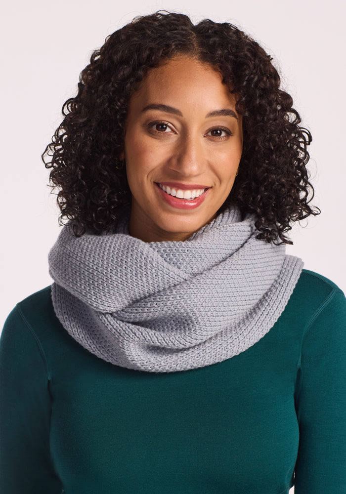 A woman with curly dark hair smiles while wearing the Woolx Noelle Infinity Scarf in Morning Fog and a dark green top, set against a plain light background.