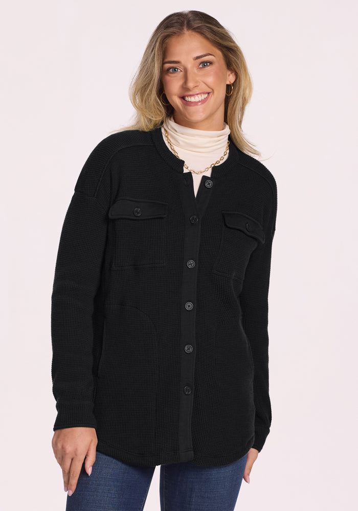 A woman with long, wavy blonde hair smiles in the Woolx Oaklynn Waffle Shacket in black, styled over a light turtleneck and blue jeans with a gold necklace, set against a plain light background. | Ariana is 5'11", wearing a size S