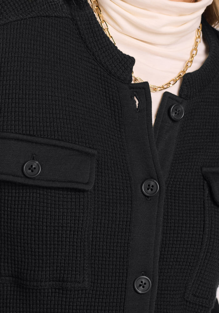 Close-up of a person wearing the Woolx Oaklynn Waffle Shacket in black with flap pockets over a cream turtleneck and a gold chain necklace. Only the lower face and upper torso are shown.