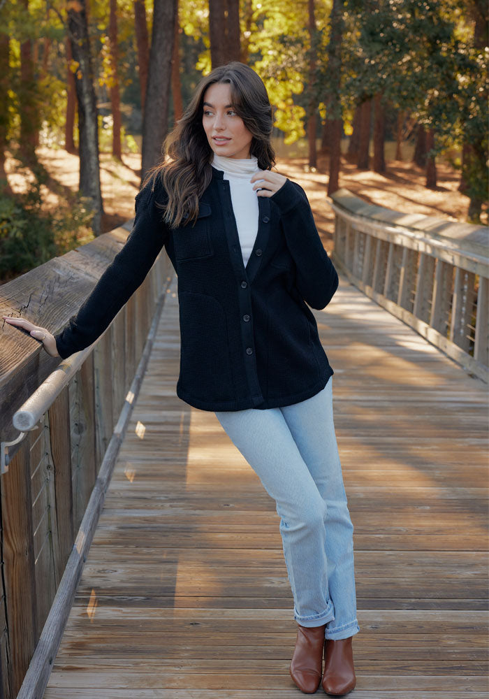 A woman in the Woolx Oaklynn Waffle Shacket in black, a white turtleneck, light blue jeans, and brown boots stands on a wooden bridge in the forest, leaning on the railing as sunlight filters through the trees behind her.