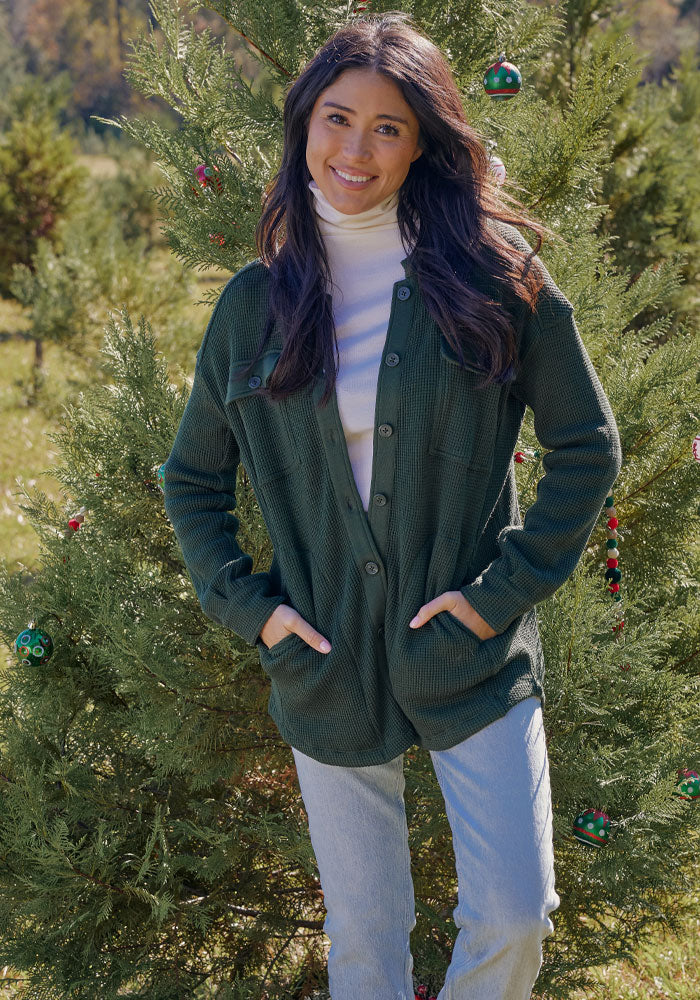 A woman with long dark hair stands outdoors in front of a decorated evergreen tree, smiling with her hands in her pockets and layering the Woolx Oaklynn Waffle Shacket in Driftwood Fern over a white turtleneck and light jeans.