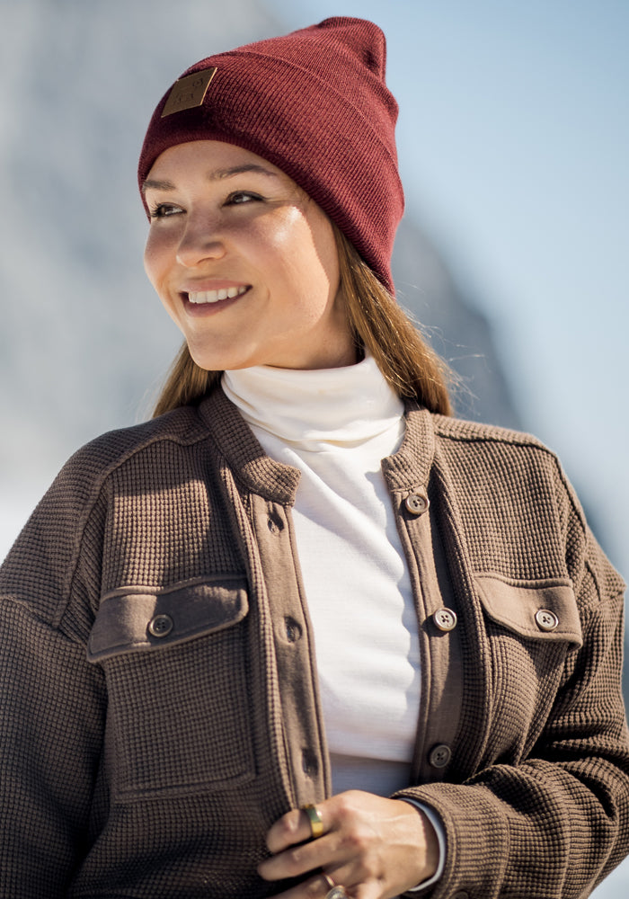 A woman smiles outdoors in a maroon beanie, white turtleneck, and the Woolx Oaklynn Waffle Shacket in Latte. The softly blurred background hints at a winter or mountain setting. The shacket is a final sale item.