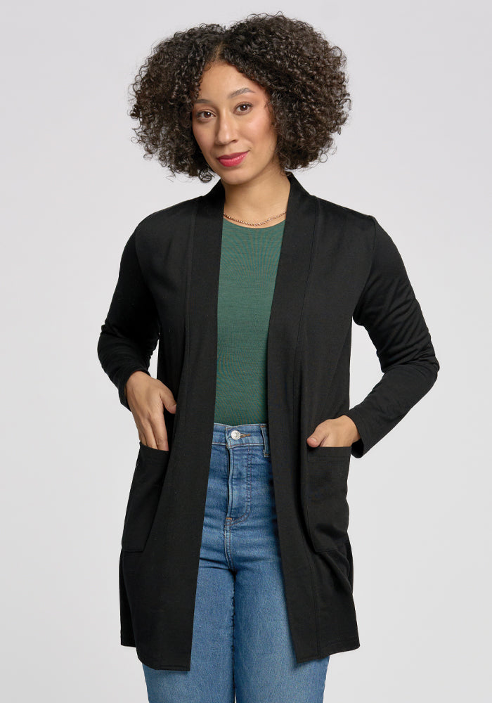 A curly-haired person wears a Woolx Paisley Cardigan in black over a green top and blue jeans. Made from lightweight Merino wool, they stand with hands in the cardigan pockets against a plain background. | Tori is 5'7", wearing a size S