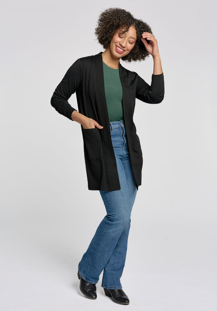 A curly-haired person smiles in a green top and Woolx's black Paisley Cardigan, which is a lightweight, mid-thigh Merino wool design. They're styled with blue jeans and black boots, one hand touching their hair as they lean slightly against a neutral backdrop.