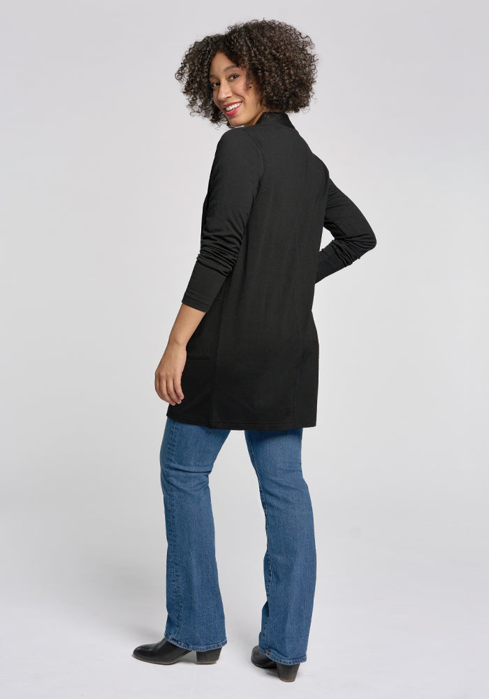 A person with curly hair smiles at the camera, wearing the stylish Paisley Cardigan by Woolx in black, along with blue jeans and black shoes, standing against a plain gray background.