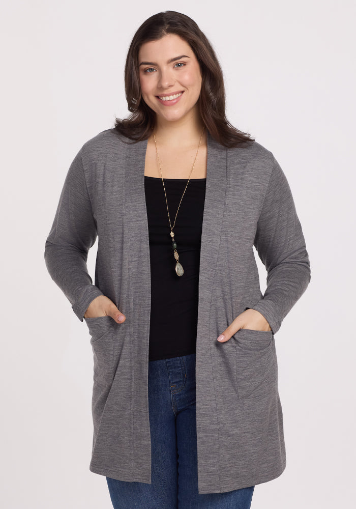 A person with long dark hair smiles, wearing a Woolx Paisley Cardigan in Graphite Heather over a black top and blue jeans. They accessorize with a long necklace, hands in pockets, standing against a plain white background, exuding feather-light comfort.