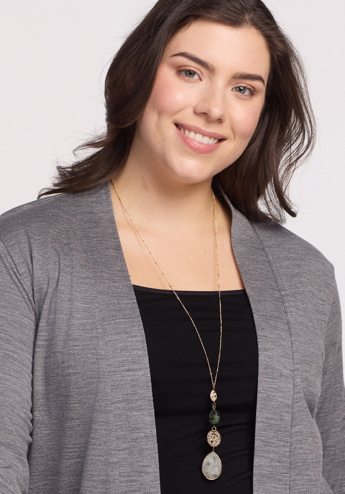 A person with shoulder-length dark hair smiles, wrapped in the feather-light comfort of Woolx’s Paisley Cardigan in Graphite Heather over a black top. They wear a long gold necklace adorned with multiple stones and a large pendant against a plain white background.