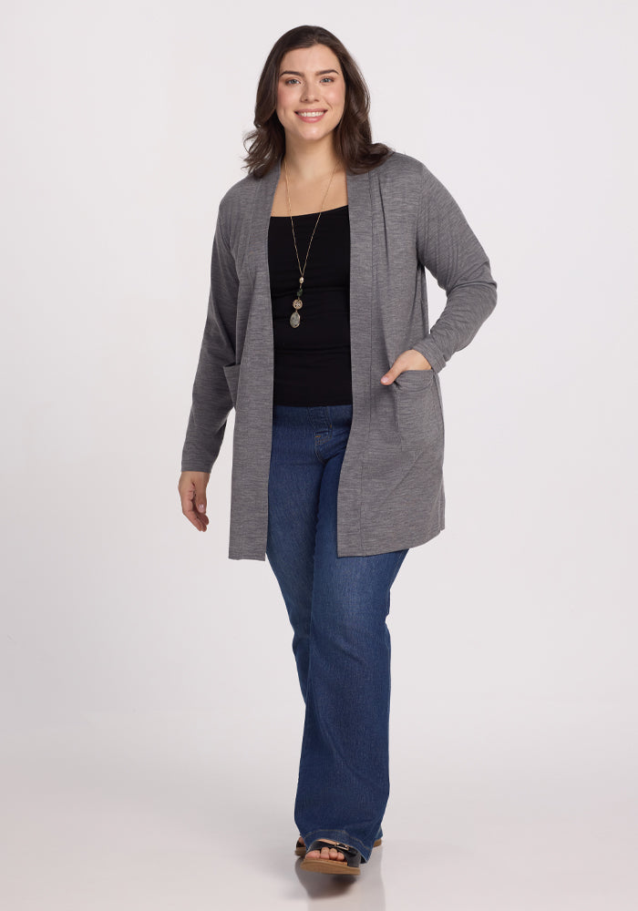 A smiling person strides forward wearing the Woolx Paisley Cardigan in Graphite Heather over a black top and blue jeans. They have long, dark hair and are accessorized with a necklace against a plain white backdrop.