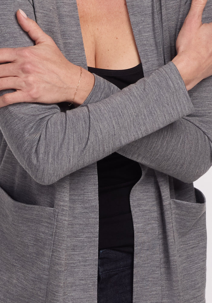 A person in a Paisley Cardigan - Graphite Heather by Woolx folds their arms, wearing a black top underneath. The women's essential piece features visible pockets, complemented by a delicate bracelet on their wrist for feather-light comfort.