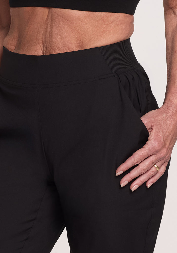 A close-up shows someone in Woolx's black Palmer Joggers, made of Merino fabric. They wear a matching black cropped top, with a gold ring on their left hand that rests on their hip. The joggers boast a gathered waistband and deep pockets against a plain white background.