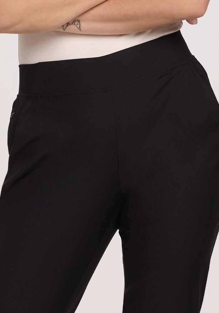 A person wearing Woolx's Palmer Joggers in black, featuring deep pockets, with a white top. Their right arm casually rests on their hip, revealing a small forearm tattoo, highlighting the pants and waist area.