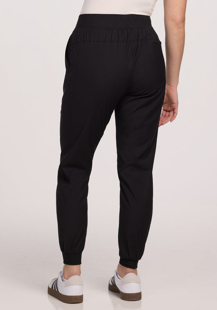 A person in Woolx's Palmer Joggers - Black and white sneakers stands with their back to the camera against a plain backdrop. Their tucked-in white shirt and joggers featuring deep pockets create a casual yet polished look.