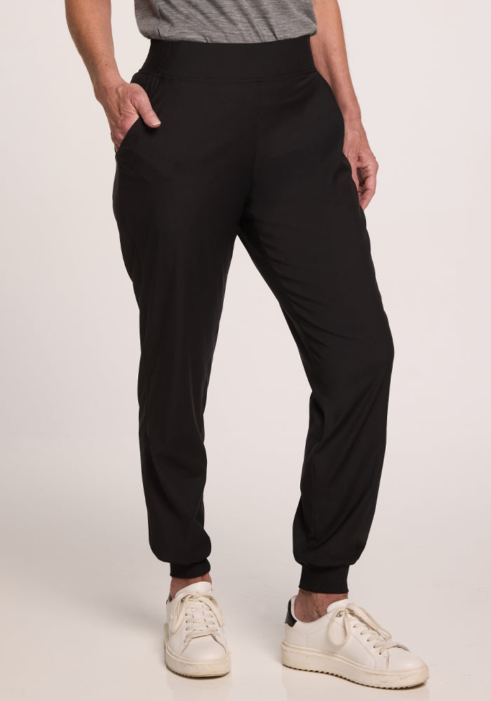 A person wearing the Palmer Joggers - Black from Woolx is pictured with one hand in their deep pocket. They're also seen wearing a gray shirt and white sneakers, standing against a plain, neutral background. | Tam is 5’8.5”, wearing a size S