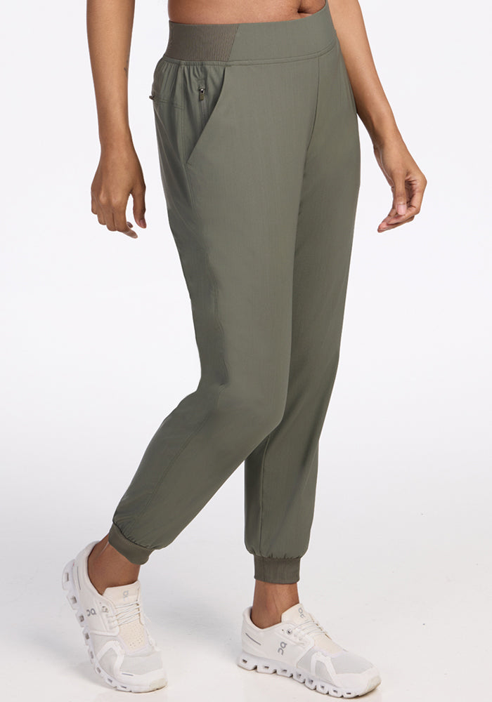 A person wearing Woolx Palmer Joggers in Sage Olive, featuring zippered pockets, and white sneakers, stands against a plain white background.