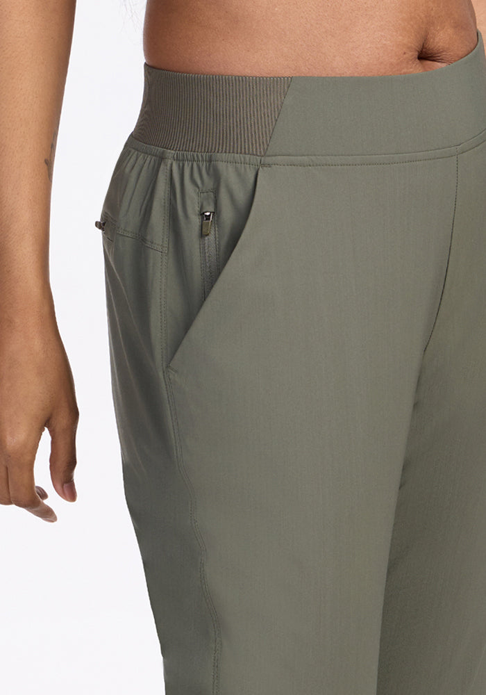 A close-up of a person wearing Woolx Palmer Joggers in Sage Olive, featuring an elastic waistband and zippered side pocket, with part of their bare torso and left hand visible.