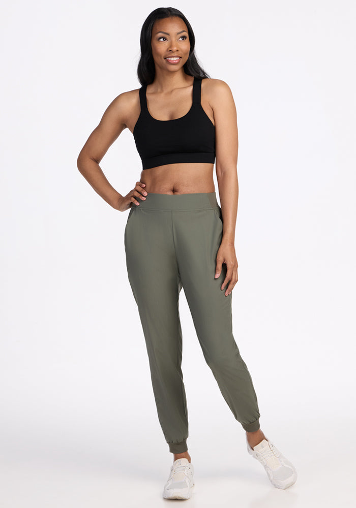 A woman smiles against a white background, wearing Woolx Palmer Joggers in Sage Olive, a black sports bra, and white sneakers. One hand is in her pocket while the other hangs by her side.