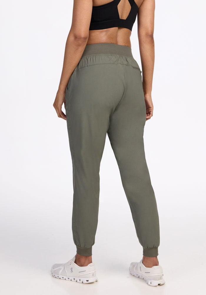 A person wearing Woolx Palmer Joggers in Sage Olive, a black sports bra, and white sneakers stands with their back to the camera against a plain white background.