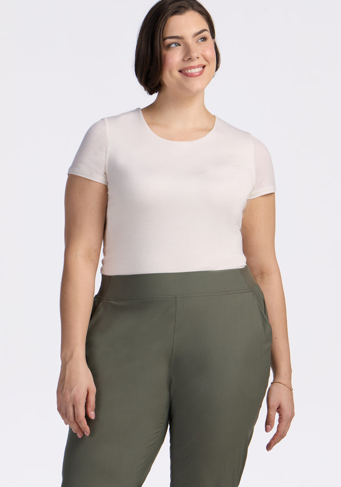 A woman with short brown hair smiles while wearing a white short-sleeve t-shirt and Woolx Palmer Joggers in Sage Olive, standing against a plain white background.