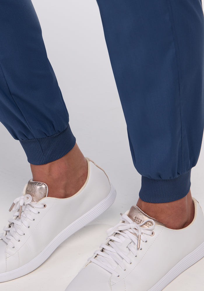 Close-up of a person wearing Woolx Palmer Joggers in Vintage Indigo with ribbed cuffs and white sneakers featuring metallic accents, standing on a light-colored surface.