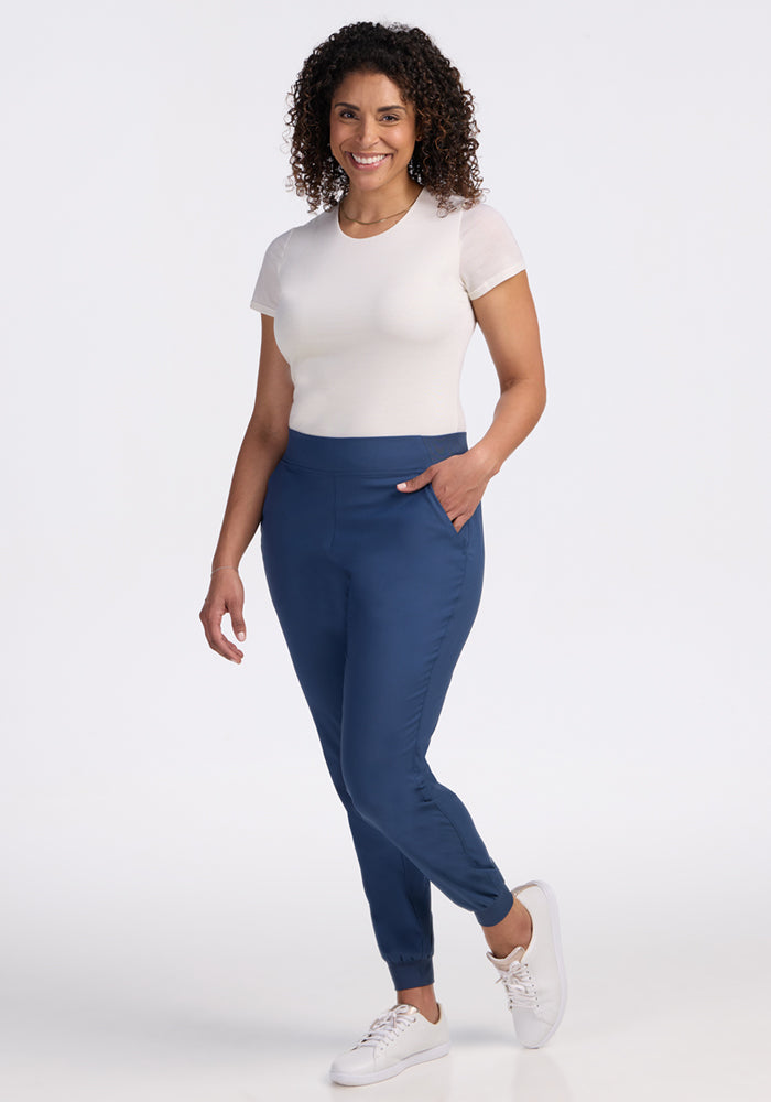 A woman with curly hair smiles, standing against a white background, wearing Woolx Palmer Joggers in Vintage Indigo and a fitted white t-shirt, with one hand in her pocket and white sneakers.