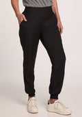 A person wearing the Palmer Joggers - Black from Woolx is pictured with one hand in their deep pocket. They're also seen wearing a gray shirt and white sneakers, standing against a plain, neutral background. | Tam is 5’8.5”, wearing a size S