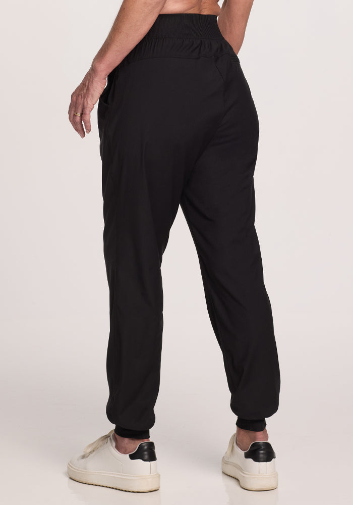 Rear view of a person wearing Woolx Palmer Joggers - Black, made from Merino fabric, paired with white sneakers featuring black soles. They stand on a plain background with hands by their sides, highlighting the joggers' deep pockets.