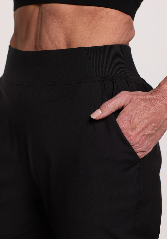 A close-up features a person in Woolx Palmer Joggers - Black, showcasing the deep pocket and waistband while wearing a black top.