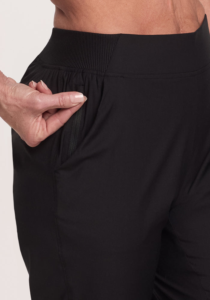 A close-up shows someone wearing Woolx Palmer Joggers - Black. They pull on the deep pocket with their thumb and index finger, highlighting the joggers' ribbed elastic waistband and pocket design.