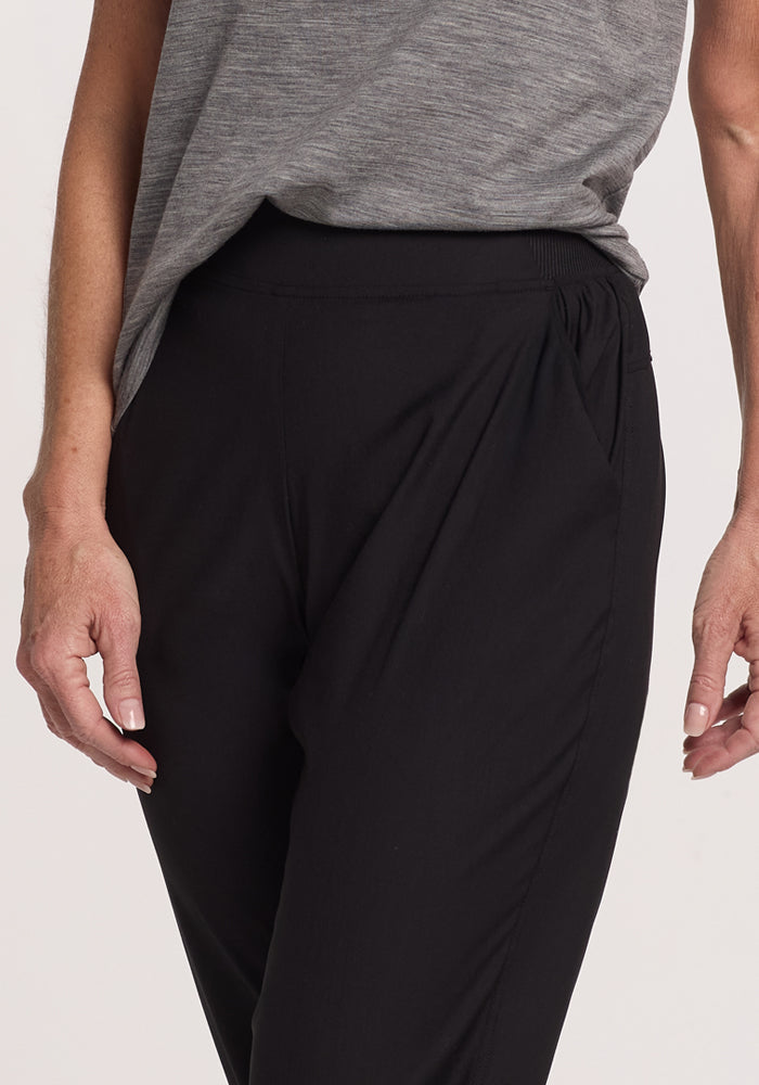 A person stands wearing Woolx Palmer Joggers in black. The joggers boast a loose fit with slight gathering at the waistband and deep pockets. One hand casually rests in a pocket, while the other arm hangs by their side, highlighting the waist and upper legs.