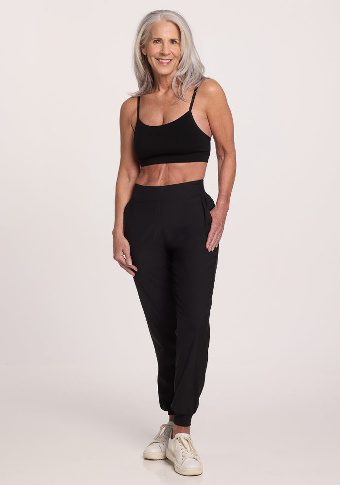 A woman with long gray hair smiles while wearing a black sports bra and Woolx's Palmer Joggers in black, made from Merino fabric. Her white sneakers complete the look against a plain, light background that highlights her fit appearance.