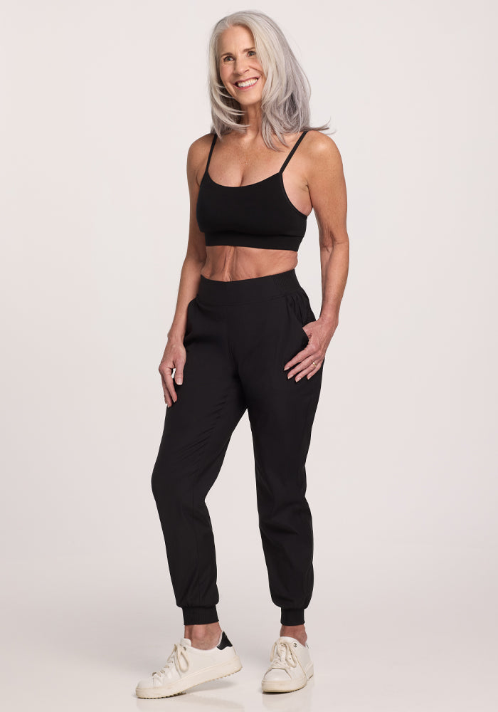 An older woman with long gray hair stands smiling in a black sports bra and Palmer Joggers by Woolx. Her joggers, featuring deep pockets, complement her white sneakers perfectly as she maintains a relaxed, confident pose against a plain white background.