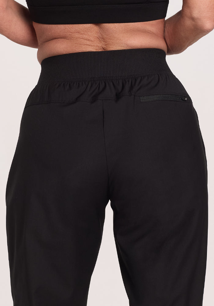 Back view of a person wearing Woolx Palmer Joggers in black, made from Merino fabric with a ribbed elastic waistband and zippered back right pocket. Paired with a black top, these joggers offer both style and comfort.