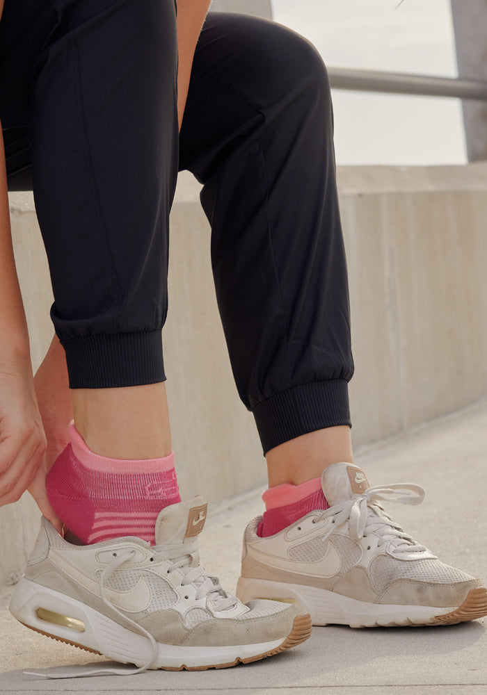 A person wearing Woolx Palmer Tall Joggers in black and white Nike sneakers with beige accents adjusts bright pink ankle socks while seated on a concrete outdoor step.